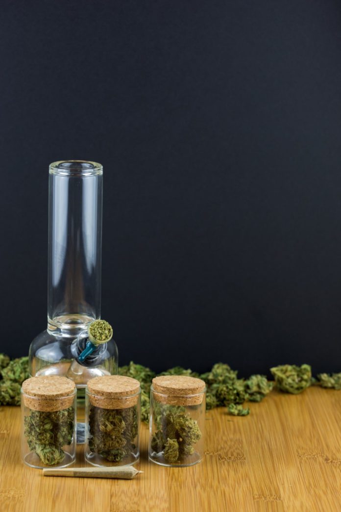 Glass jars filled with medical marijuana in a row with loose marijuana, glass bong, and joint on a bamboo table with black background in portrait