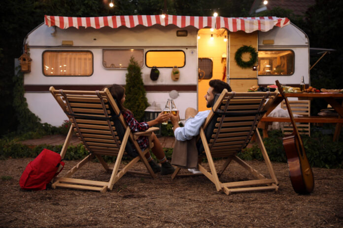 Young couple toasting with bottles of beer near trailer. Camping season